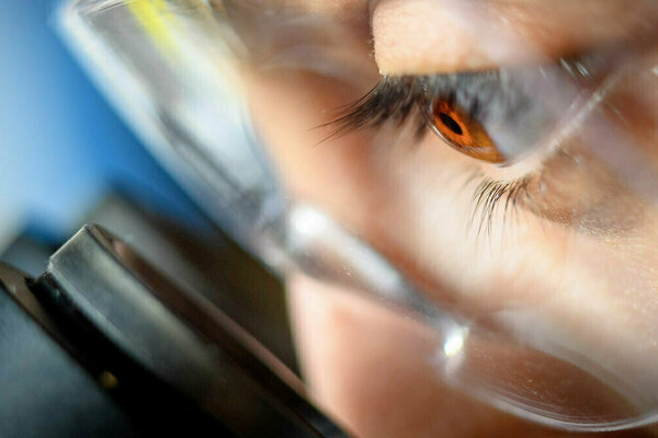 Close-up of a person's hazel eye looking intently through clear glasses at a Zeiss microscope.