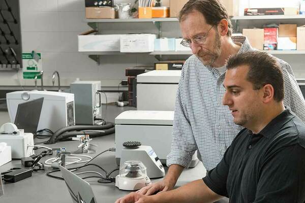 Two scientists, one with a beard and glasses, intently view a laptop in a lab with centrifuges and other equipment.