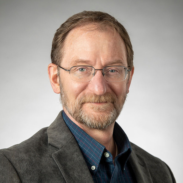 A man with light brown hair, beard, and glasses smiles, wearing a dark grey corduroy jacket and blue plaid shirt.