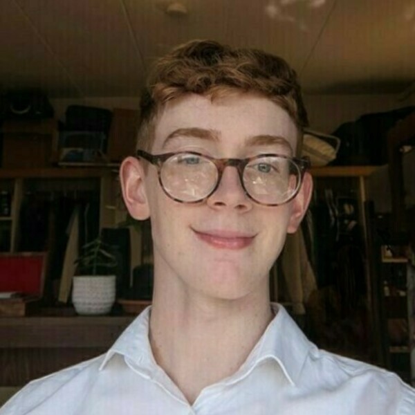 Young man with curly red hair and tortoiseshell glasses smiling. He wears a white collared shirt.