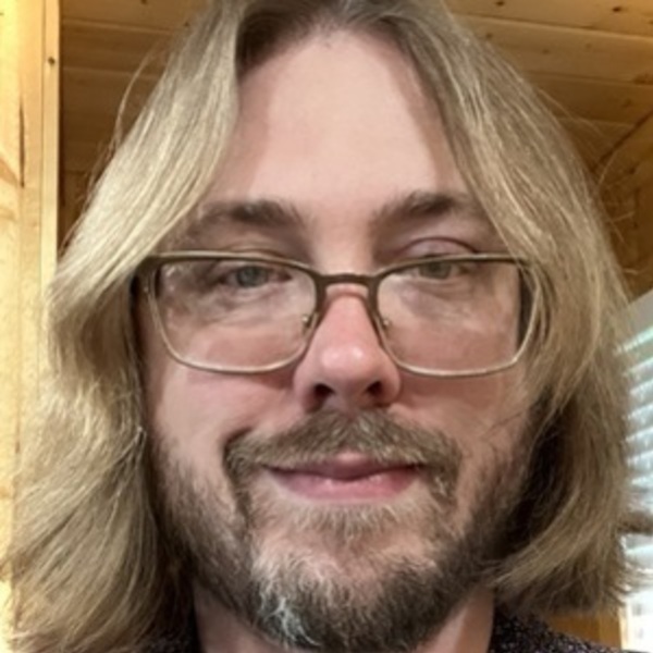 Man with blonde hair, beard, and glasses smiles subtly in a patterned shirt.