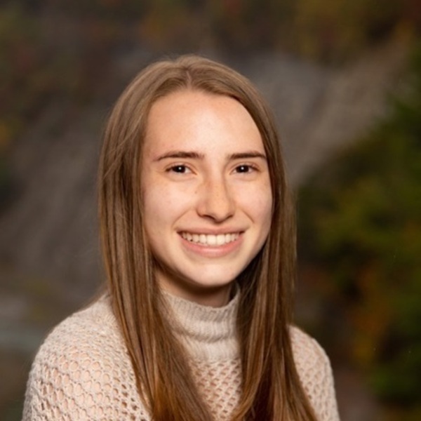 Smiling young woman with long light brown hair wears a cream knit turtleneck against a blurry natural background.