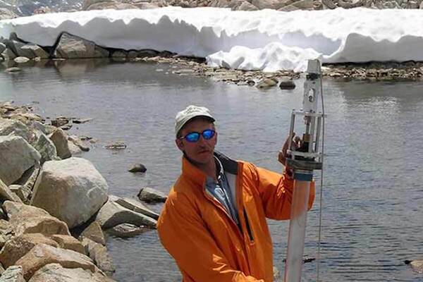 Man in orange jacket holds a clear water sampler by a rocky alpine lake and snowy peaks against a blue cloudy sky.