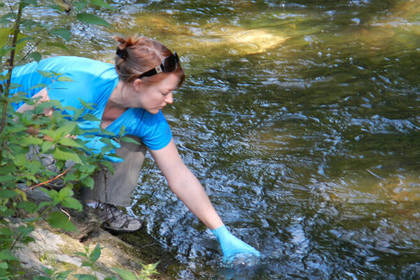 Red-haired woman in blue shirt, tan pants, and blue glove leans over a rocky bank, sampling water from a stream.